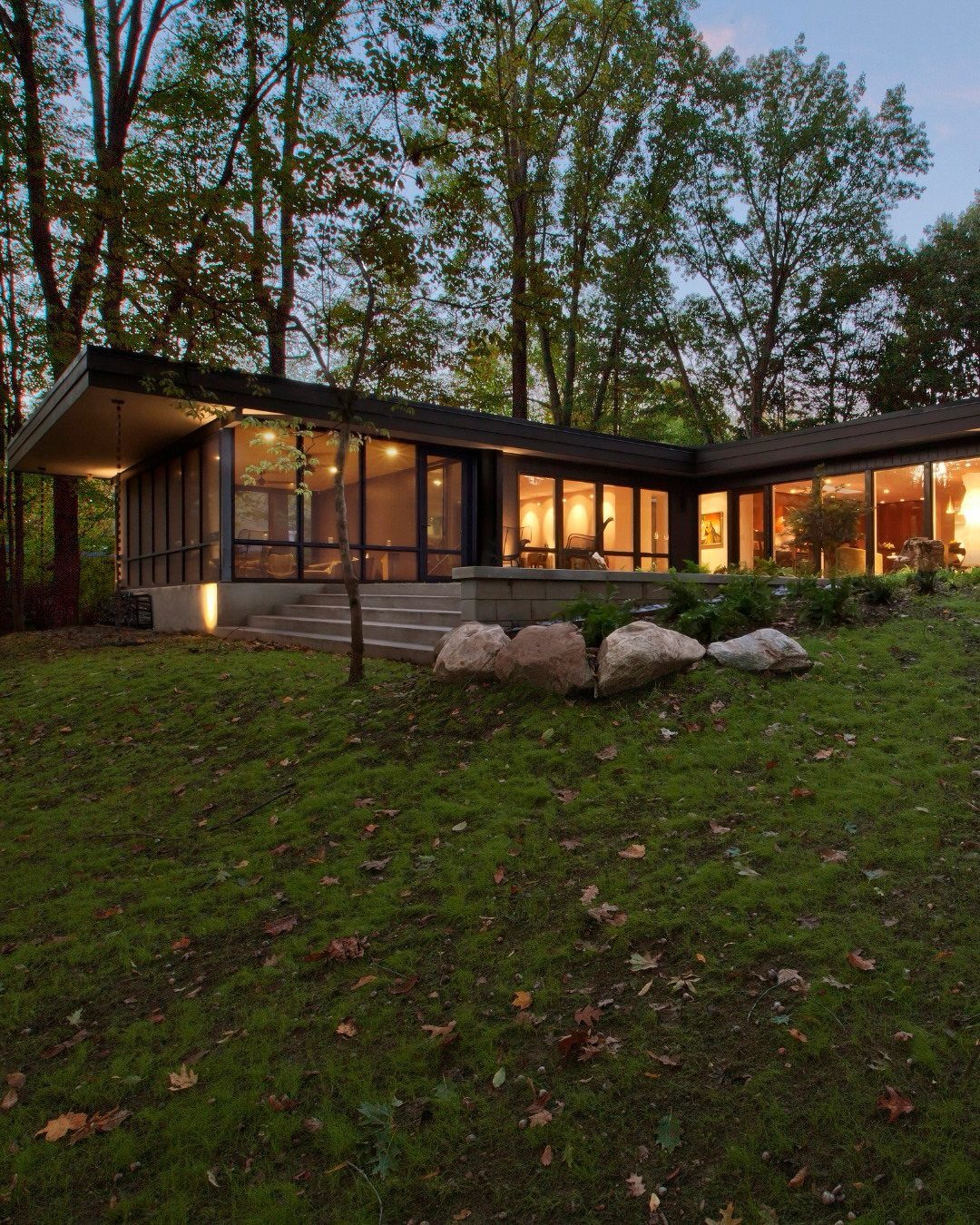 Mid-century modern house with large floor-to-ceiling glass windows surrounded by lush green trees.