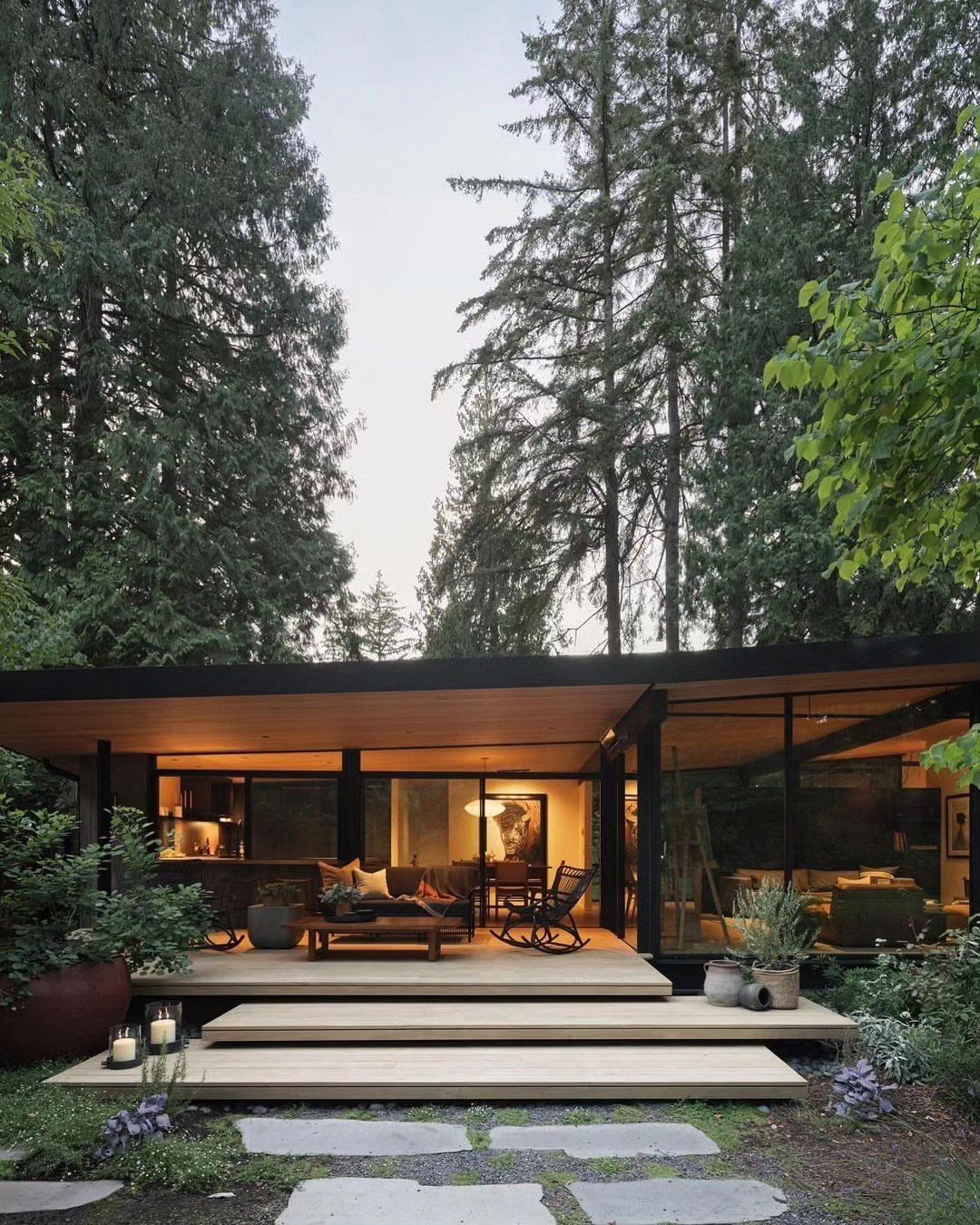 Mid-century modern house with glass walls and wooden deck surrounded by tall trees.