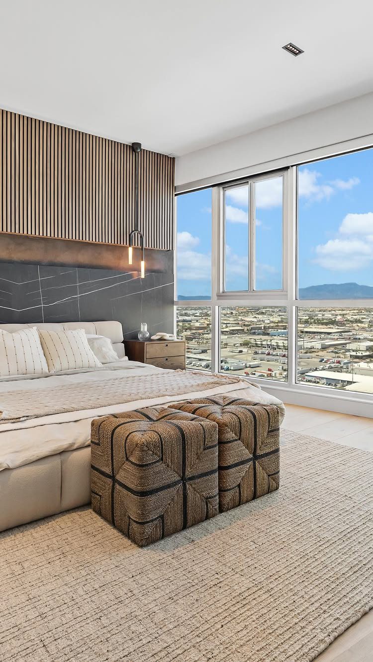 Modern bedroom with woven ottomans, beige rug, and large city view window.