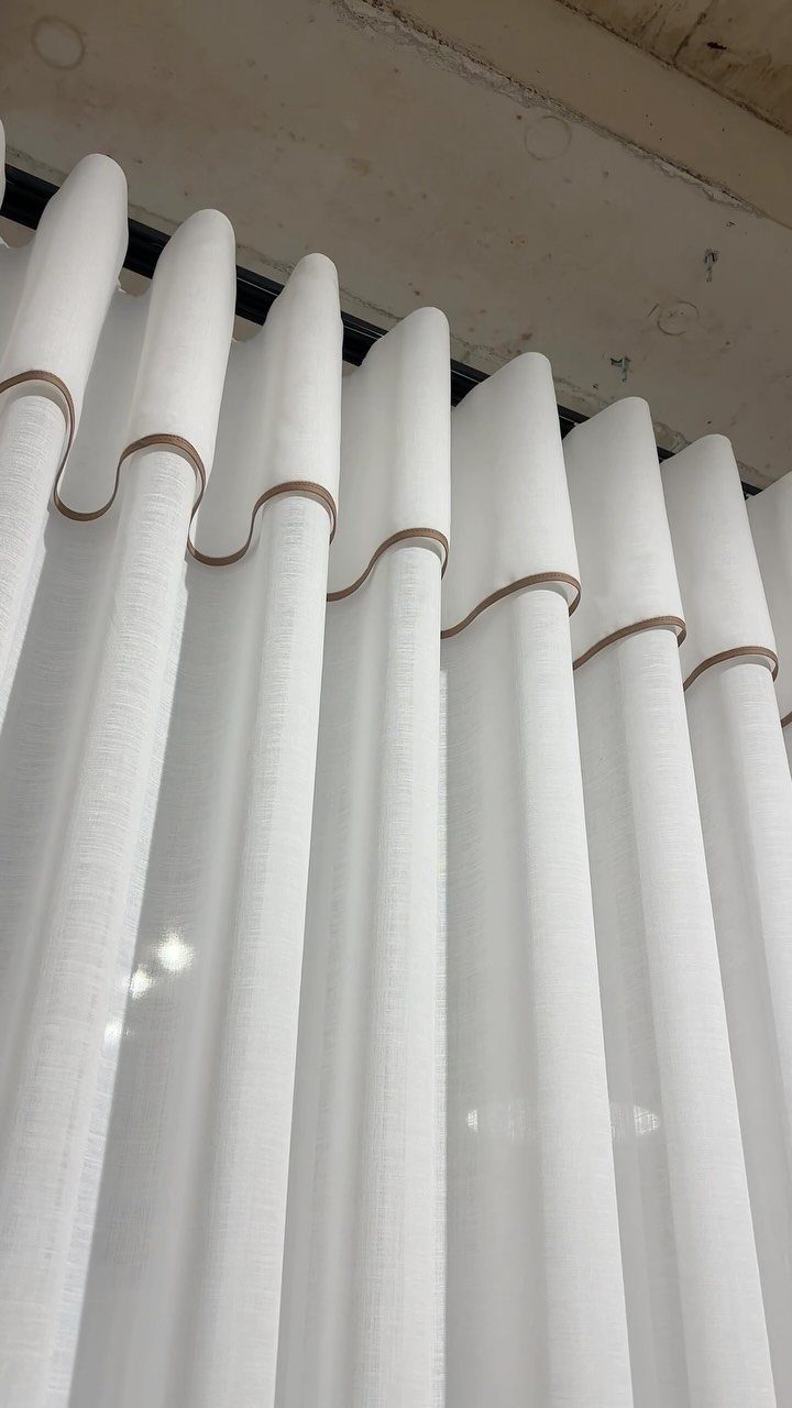 White curtains with brown piping hanging in folds from a dark rod.