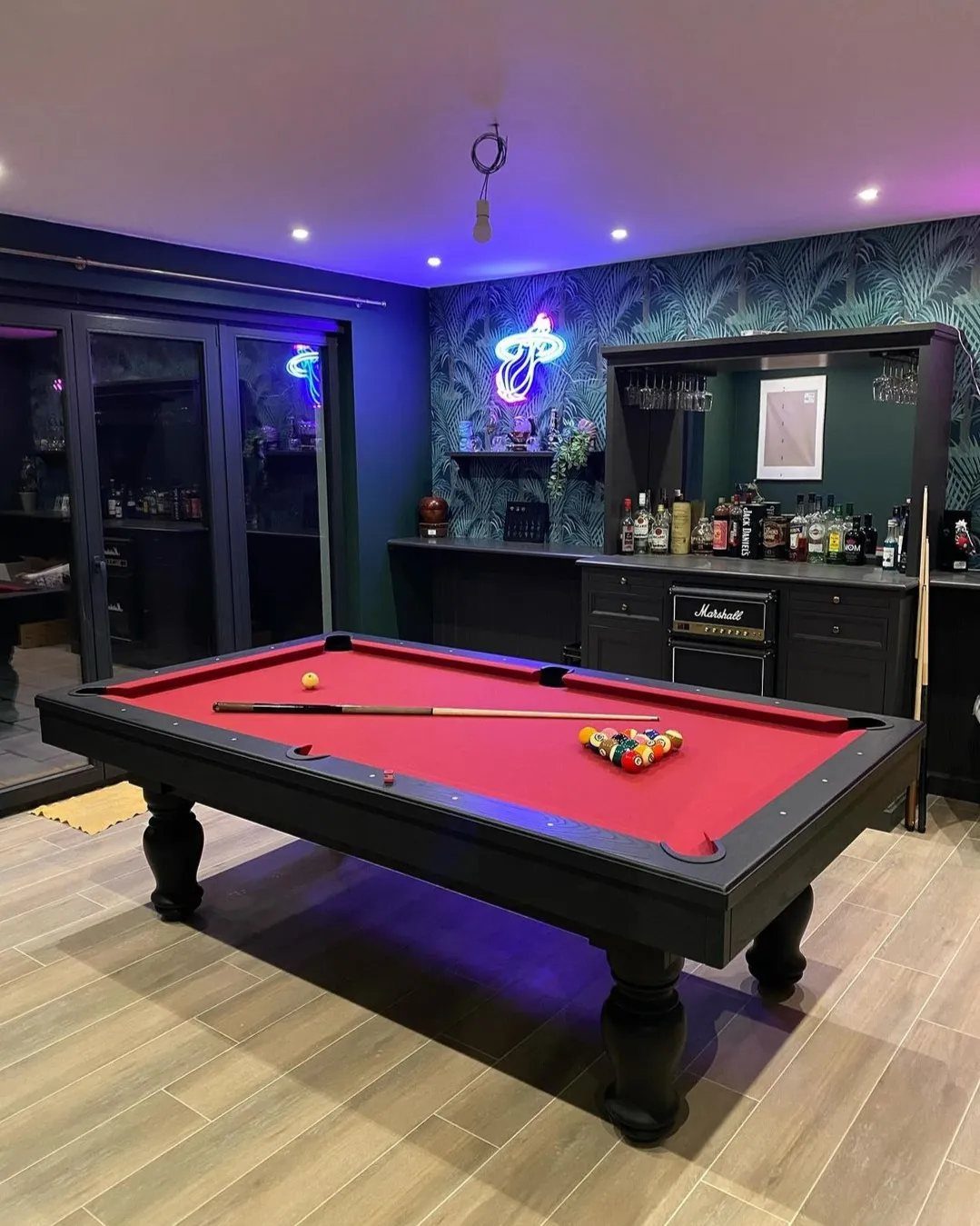 Pool table with red felt in a room with dark teal wallpaper and bar.