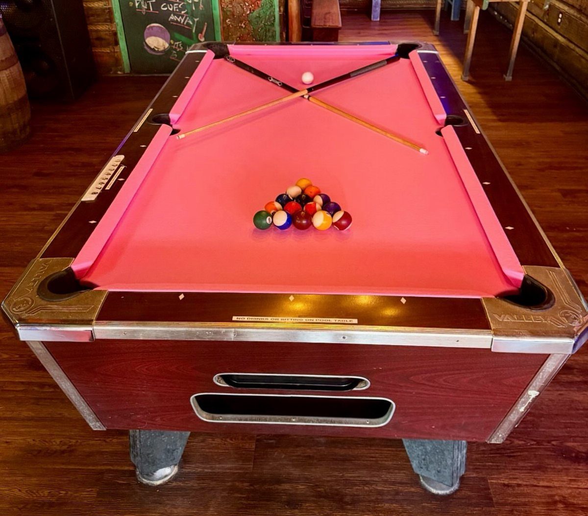 Billiard table with bright pink felt, colorful balls, and crossing cues on hardwood flooring