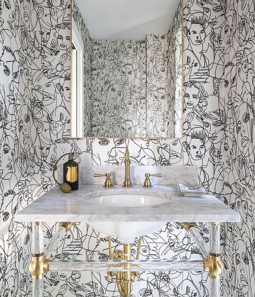 Marble sink with gold fixtures against a black and white face mural wallpaper.