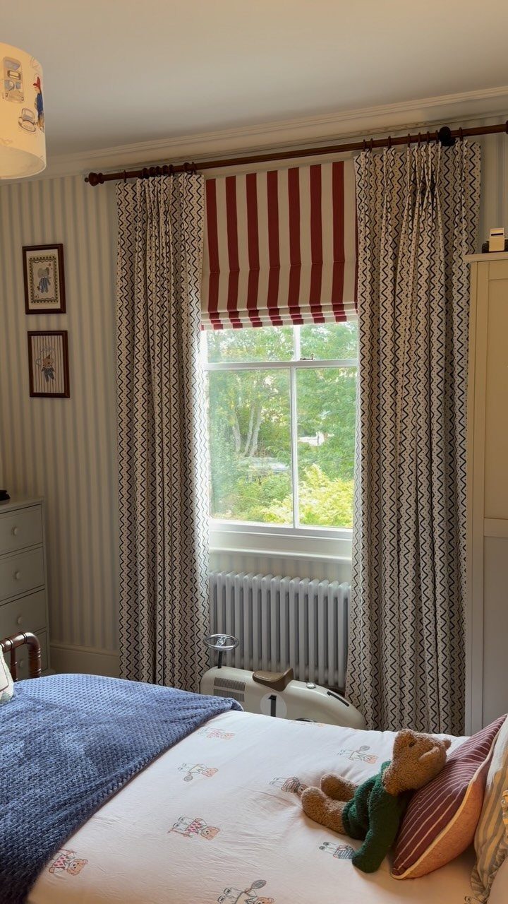 Red striped Roman blinds and patterned curtains in a child's bedroom with teddy bear.