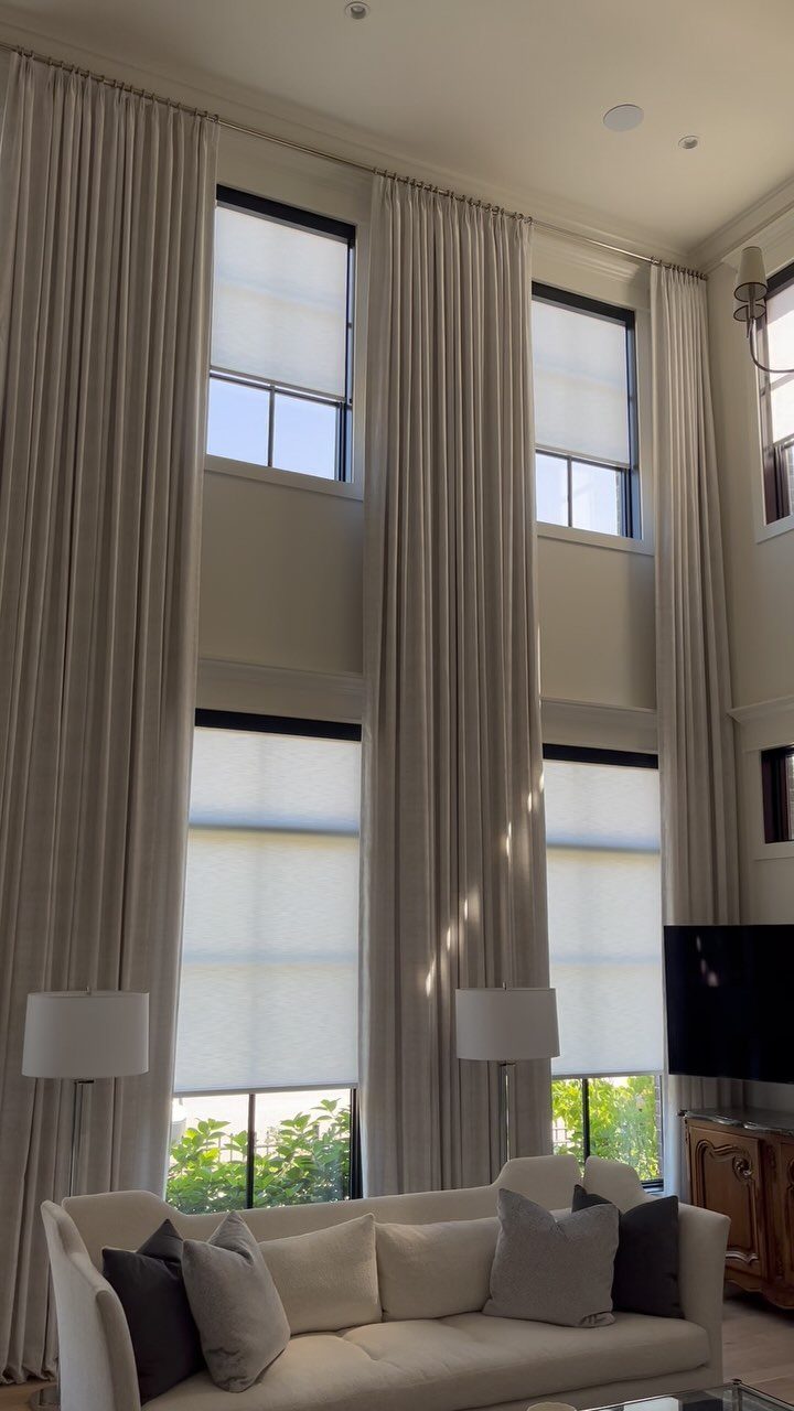 Cream curtains hanging from a high rod above windows with white roller blinds.