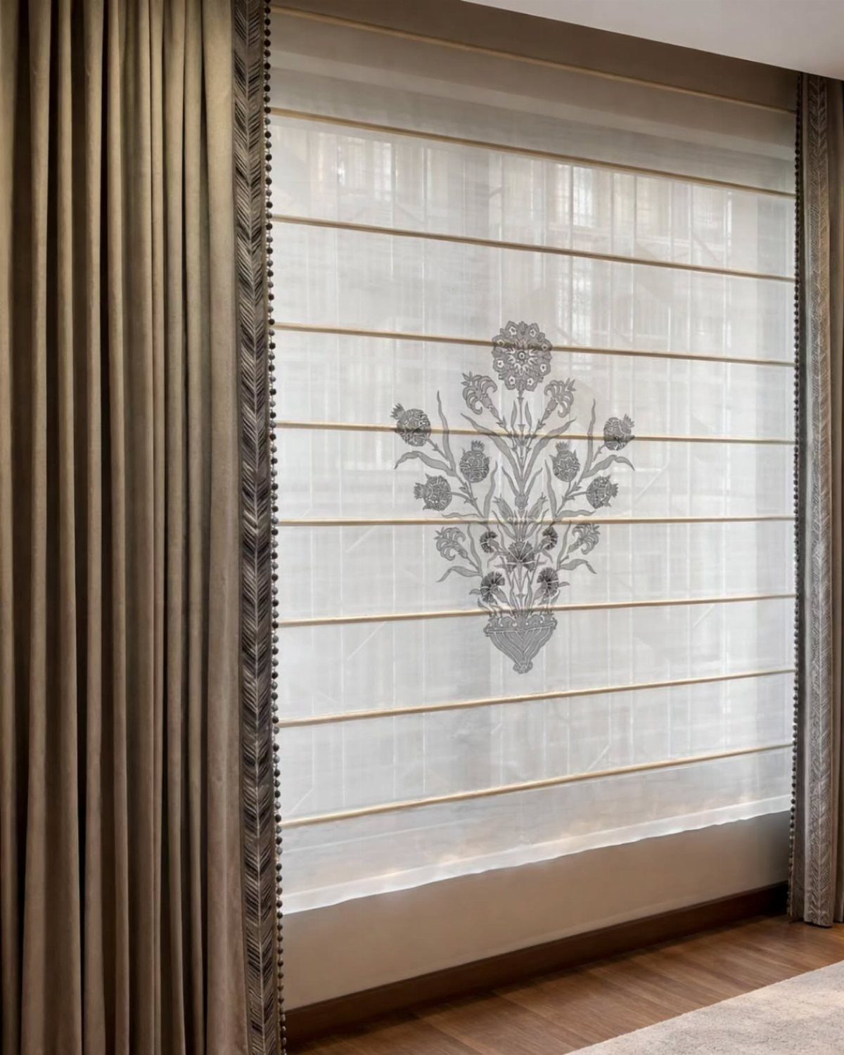 Taupe curtains framing white Roman blinds with a grey floral design on a window.