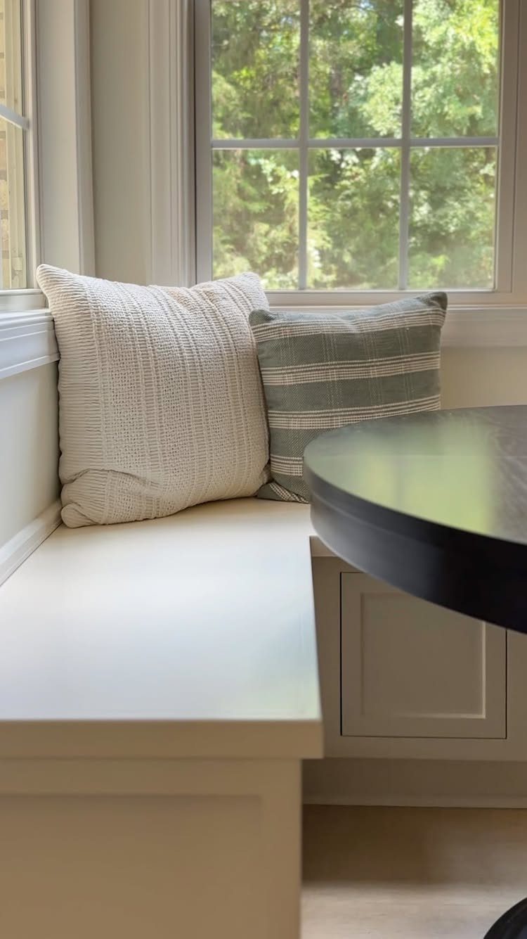 White built-in dining bench with decorative pillows next to a dark round table.
