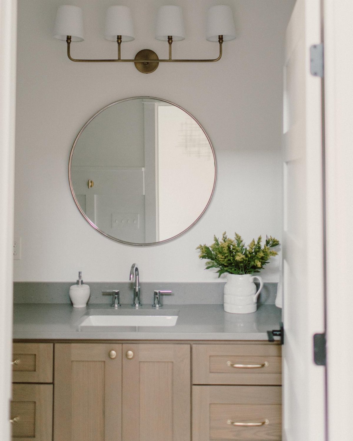 Bathroom vanity with brass light fixture, round mirror, and light wood cabinets.