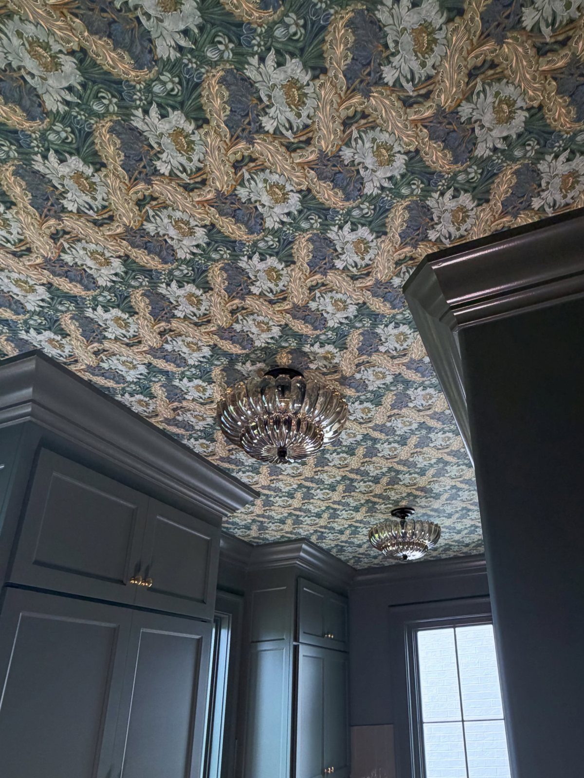 Floral patterned wallpaper covering a ceiling above dark grey cabinets and crystal chandeliers