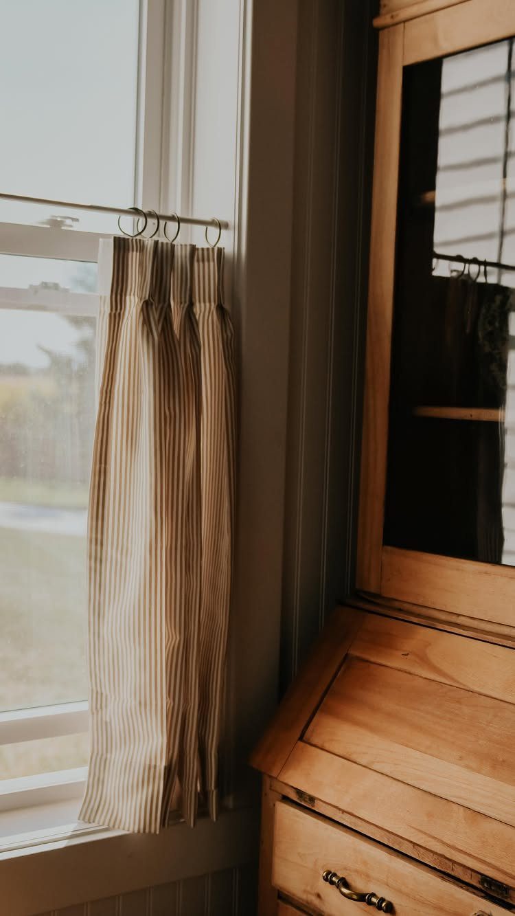 Striped café curtains hanging on a window next to a wooden secretary desk.
