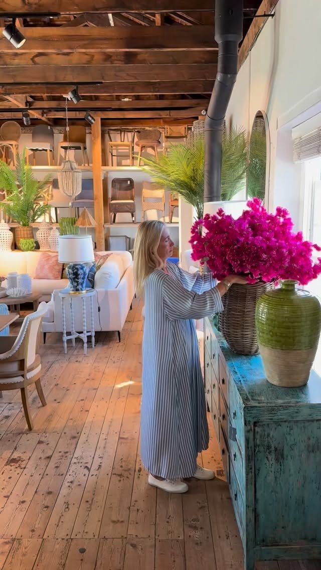 Woman arranging pink flowers on a weathered blue sideboard in a rustic room.
