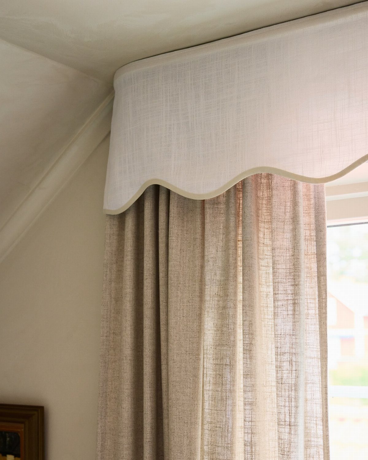 White scalloped valance hanging over beige semi-sheer linen curtains by a window.