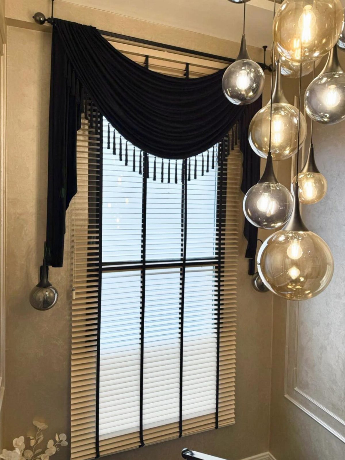 Black draped valance with fringe over light-colored window blinds and glass pendant lights.