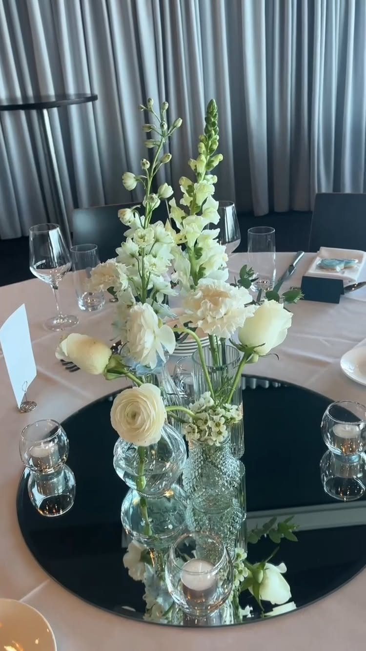 Cluster of clear glass vases with white flowers on a black mirrored tray.