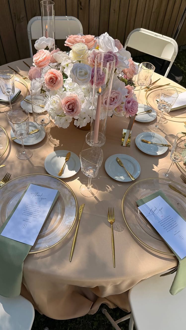 Round dinner table with beige linens, gold chargers, and pink rose centerpiece.