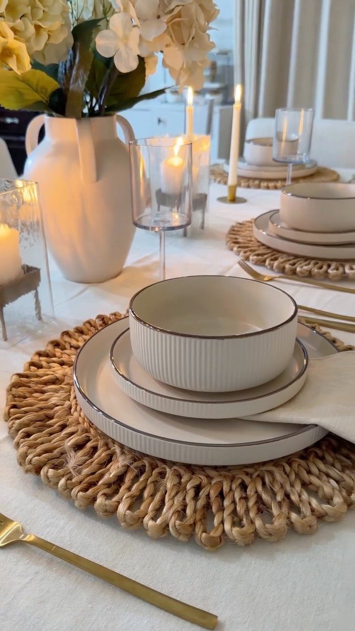 Set dinner table with white dishes, woven placemats, gold cutlery, and cream flowers.
