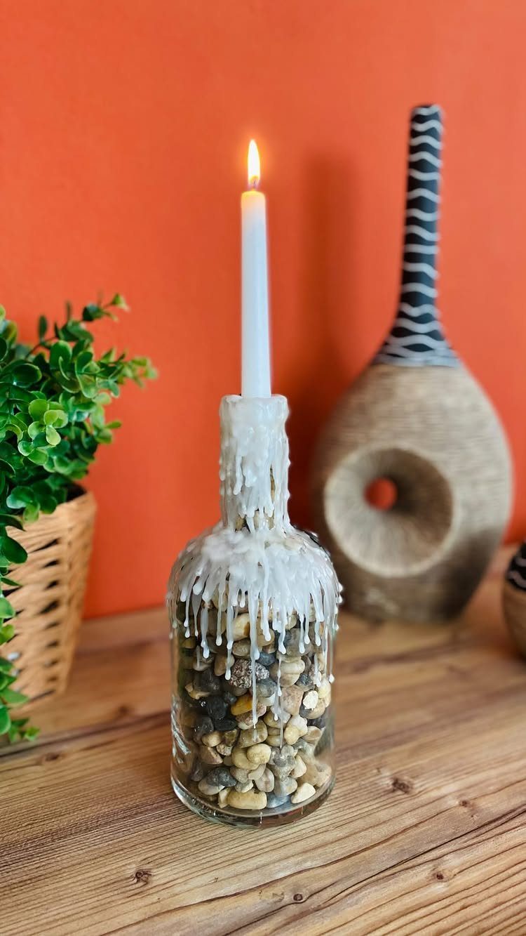 Lit white taper candle in a glass bottle filled with pebbles and melted wax.