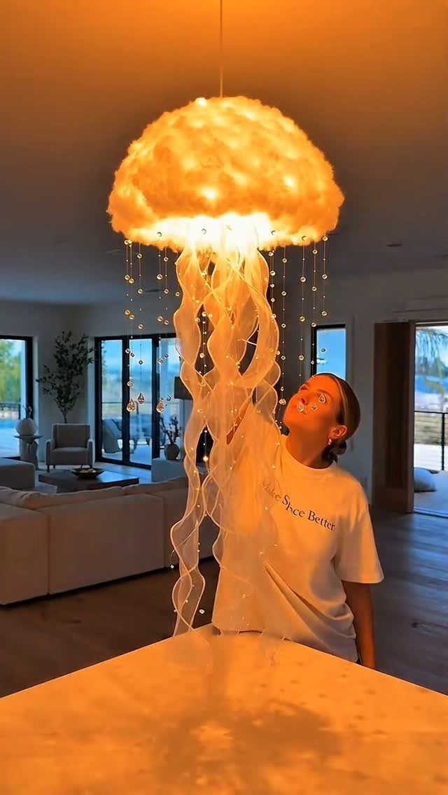 Woman looking up at a glowing jellyfish-shaped cloud lamp with flowing fabric tentacles.