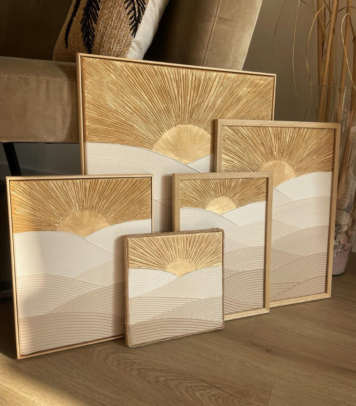 Five framed textured artworks with golden sunbursts and beige landscapes leaning against a sofa.
