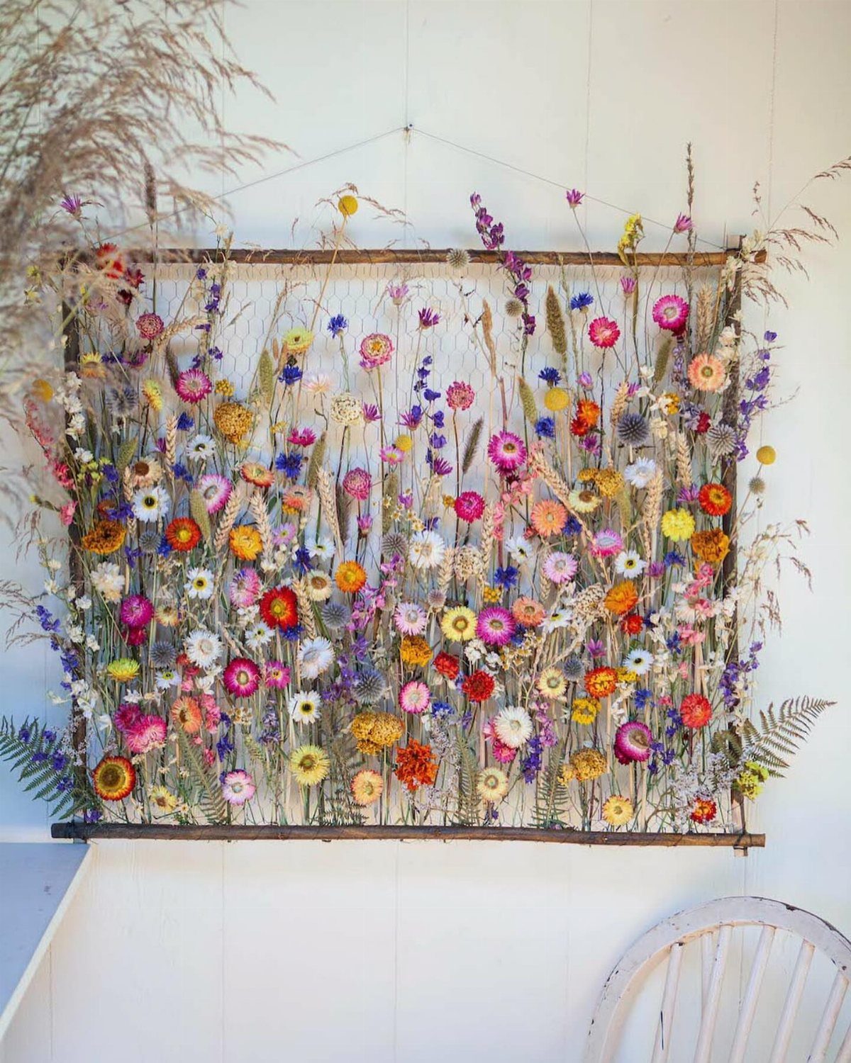 Dried colorful wildflowers arranged within a wooden frame hanging on a white wall.