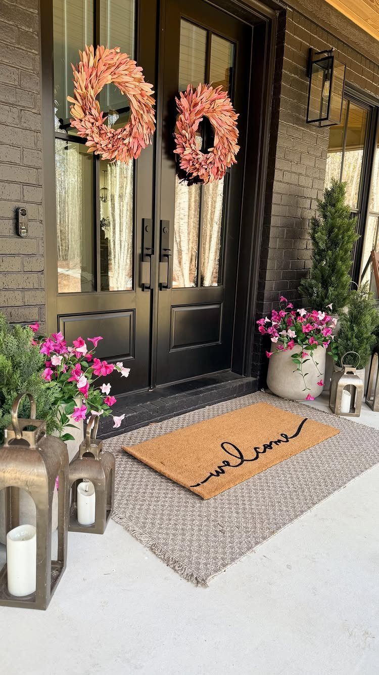 Layered doormats with a welcome mat on a grey rug before black doors.