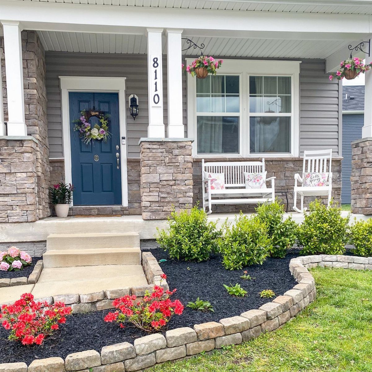 Grey house porch with blue door, white furniture, and pink hanging flower baskets.