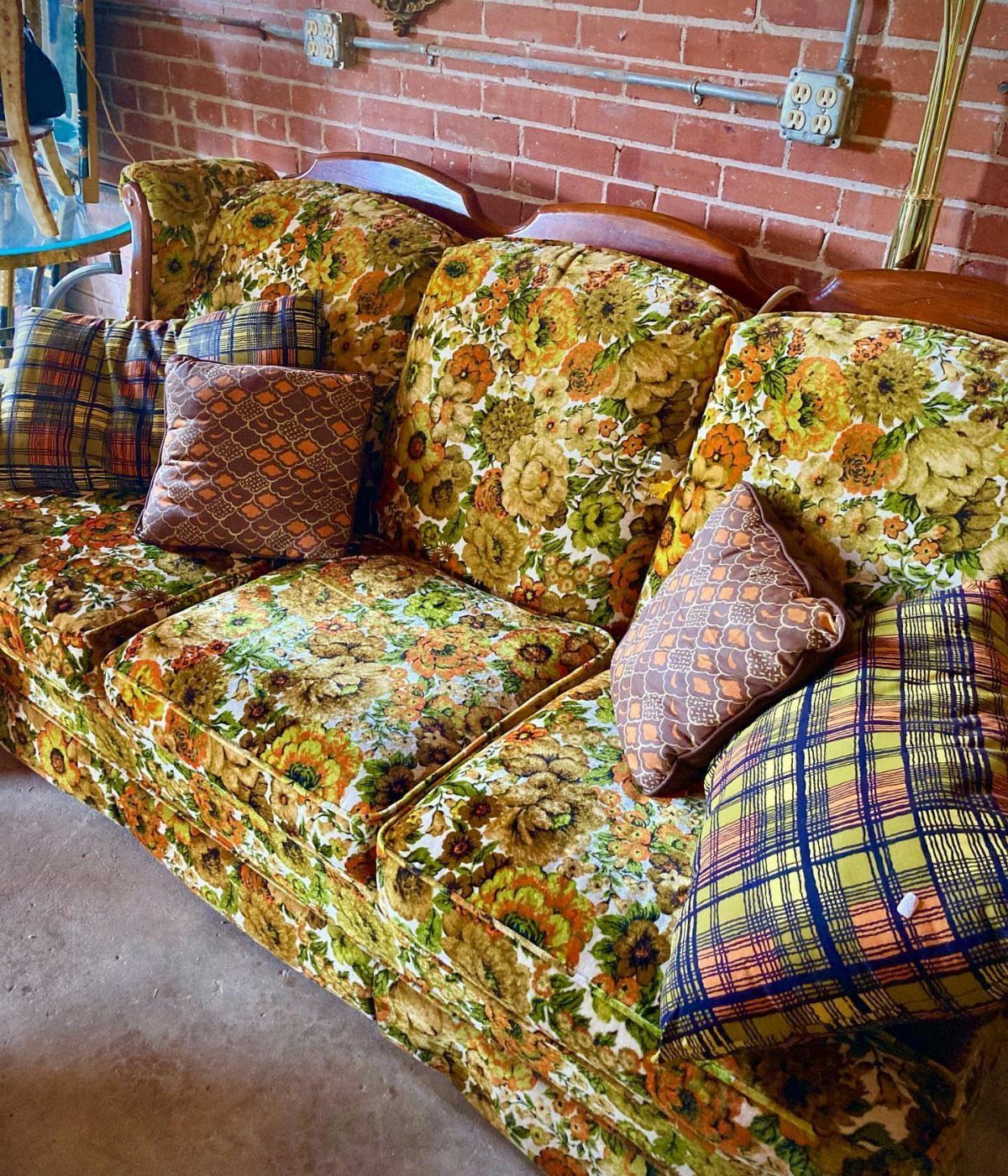 Floral patterned vintage sofa with colorful plaid pillows against a red brick wall.