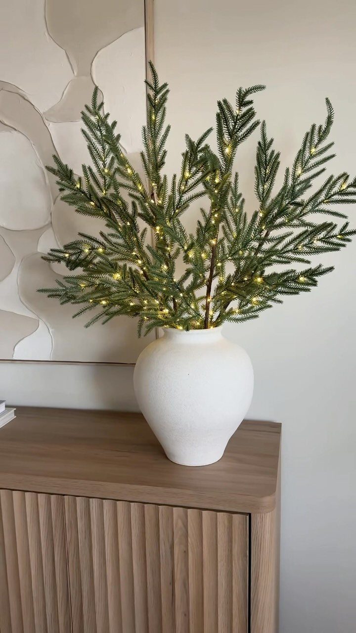 Lighted pine branches in a white vase on a wooden cabinet.