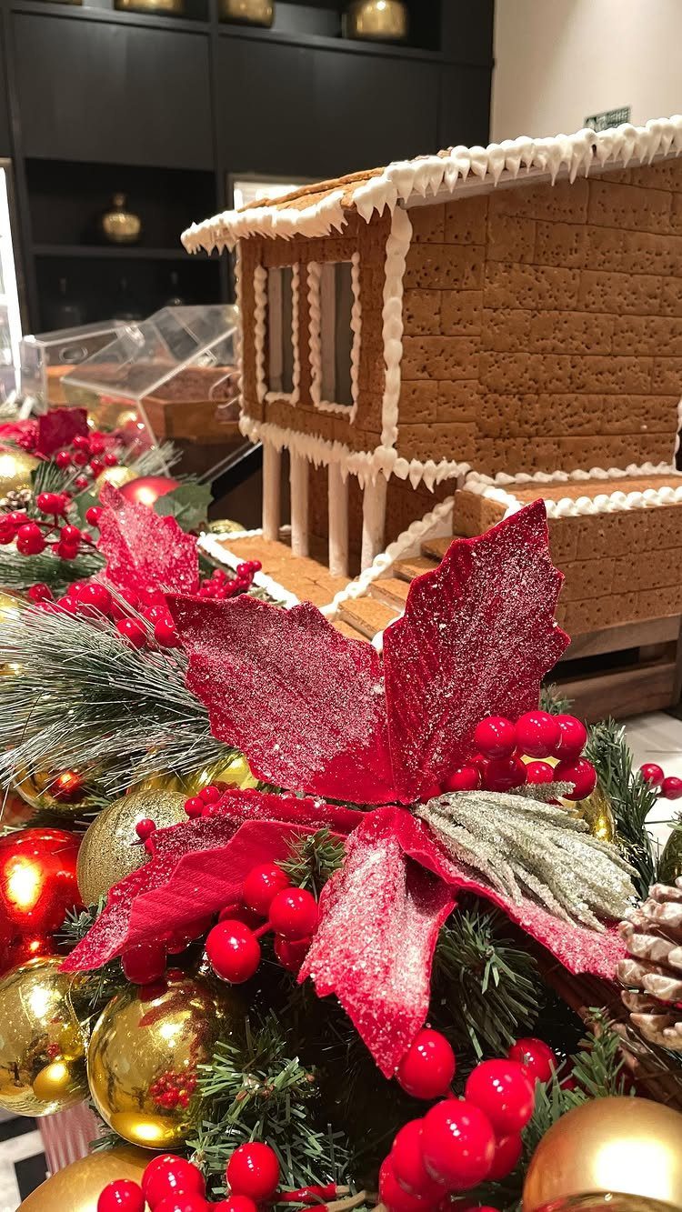 Gingerbread house behind red poinsettias, gold ornaments, and green pine needles.
