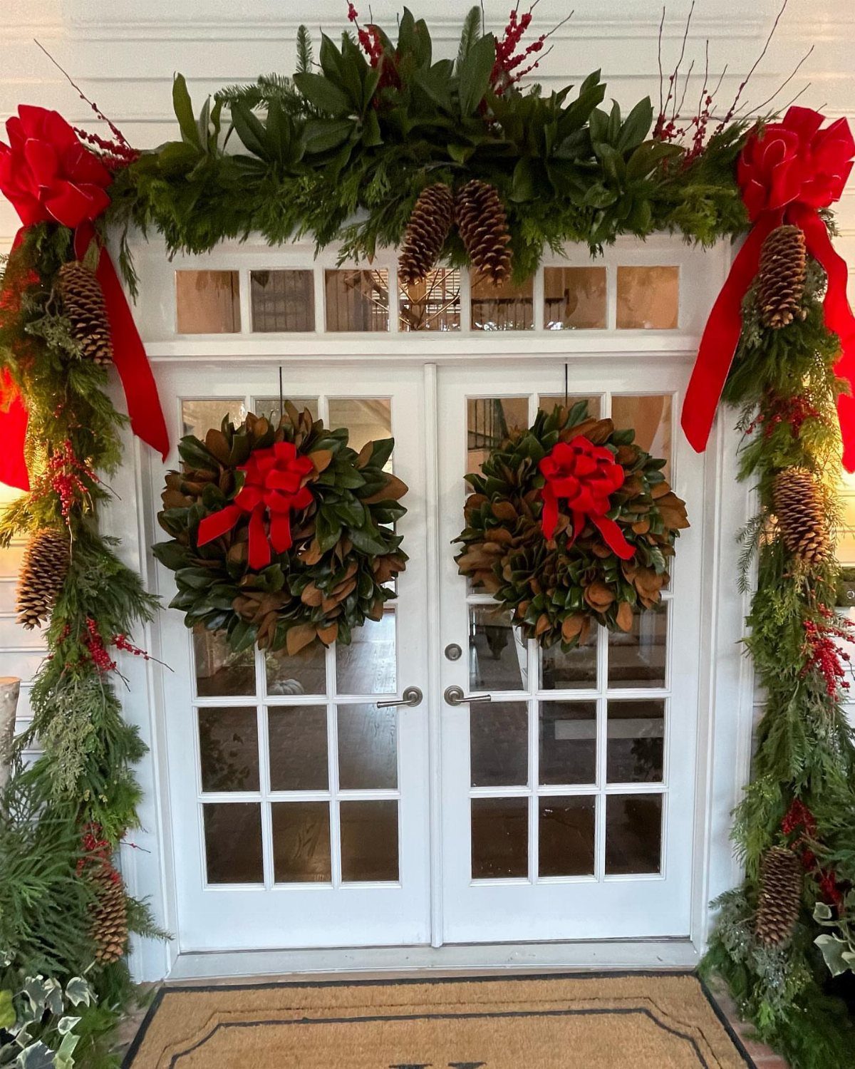 Two magnolia leaf wreaths with red bows hanging on white double doors.