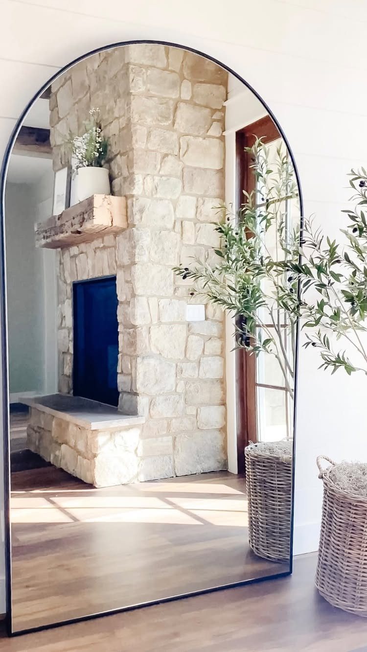 Large arched floor mirror reflecting a stone fireplace in a bright living room