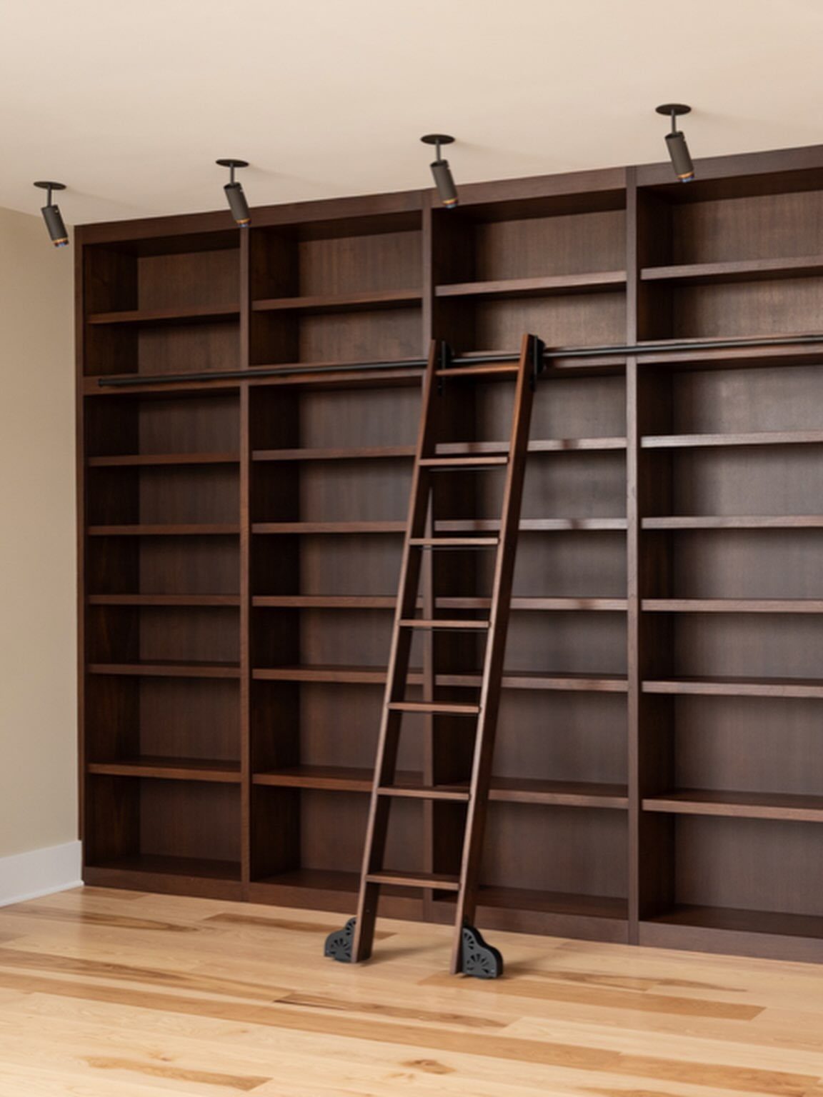 Dark brown built-in bookshelves with a matching rolling library ladder and ceiling spotlights.
