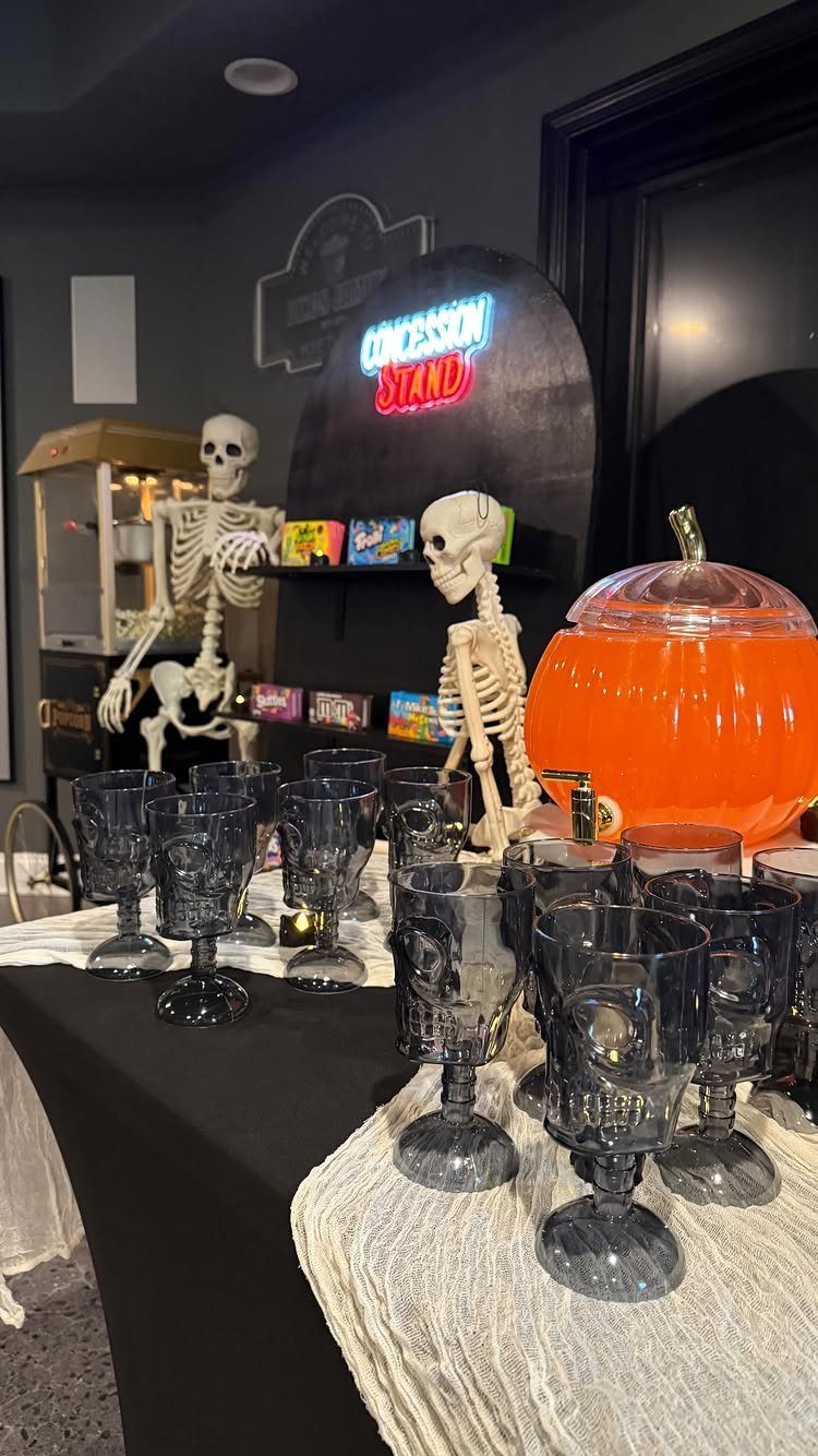 Halloween concession stand with skeletons, popcorn machine, and skull-shaped glasses on black table.