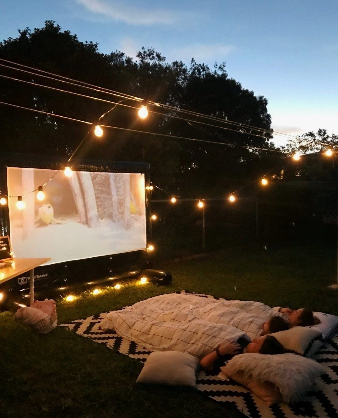 Outdoor movie projector screen with string lights and people lounging on a rug.