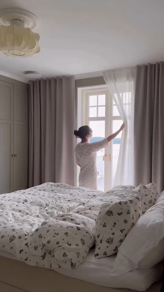 Woman opening sheer white curtains next to grey blackout curtains in a bedroom