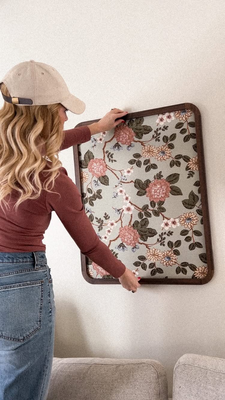 Woman hanging a large framed floral fabric art piece on a white wall.