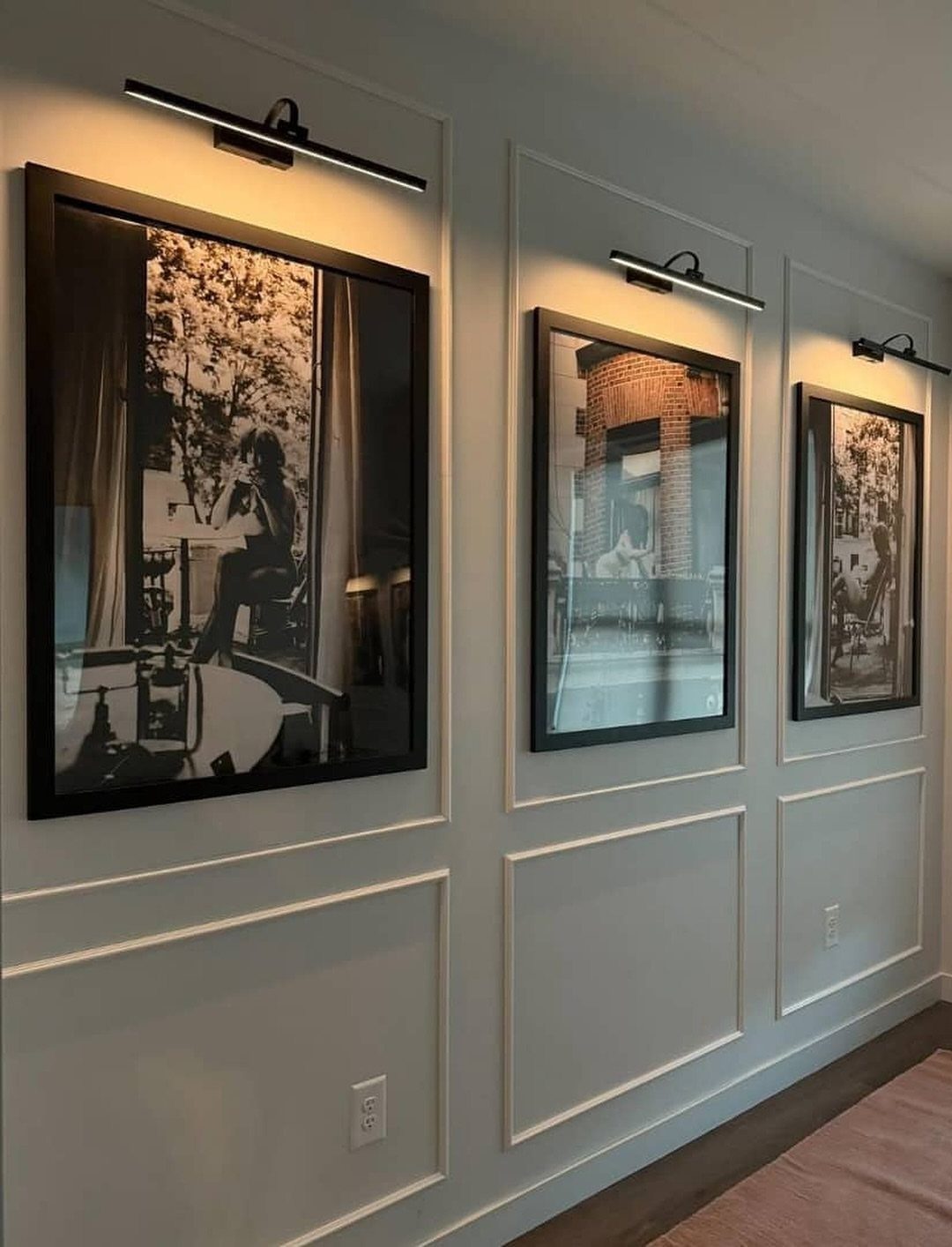 Three black-framed photographs on a white paneled wall with individual overhead picture lights.