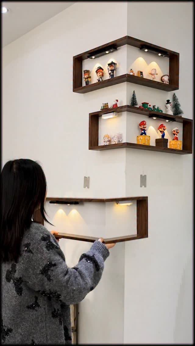 Woman installing brown L-shaped corner shelves with lighting and figurines on white walls