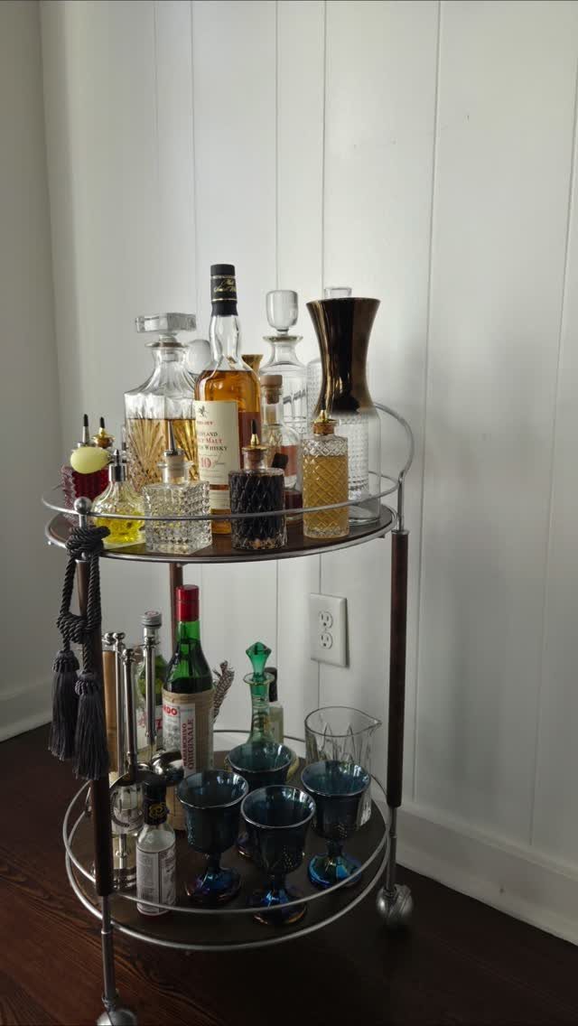 Metal bar cart with liquor bottles and blue glasses against white paneled wall