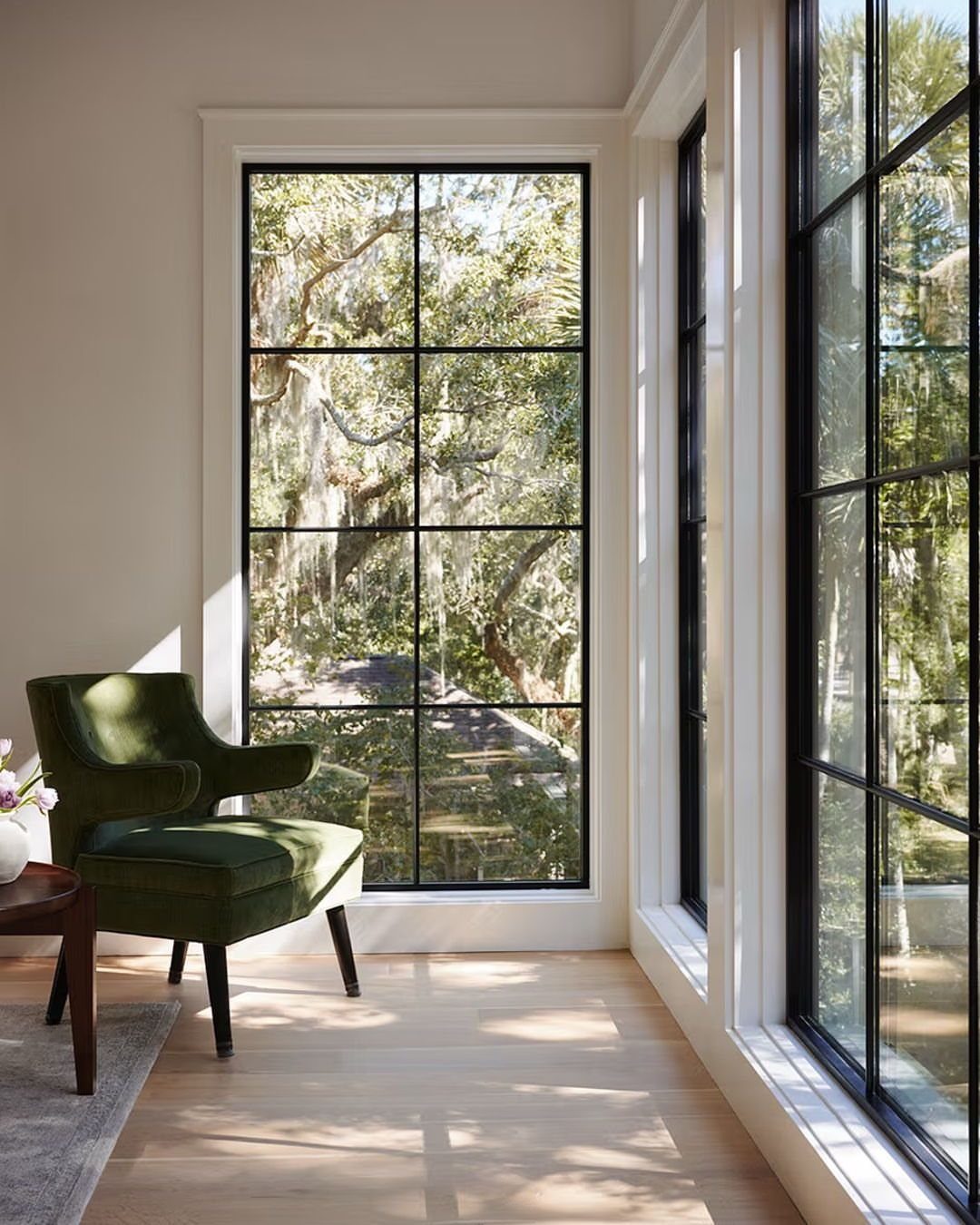Large black-framed grid windows letting in natural light next to a green velvet armchair.