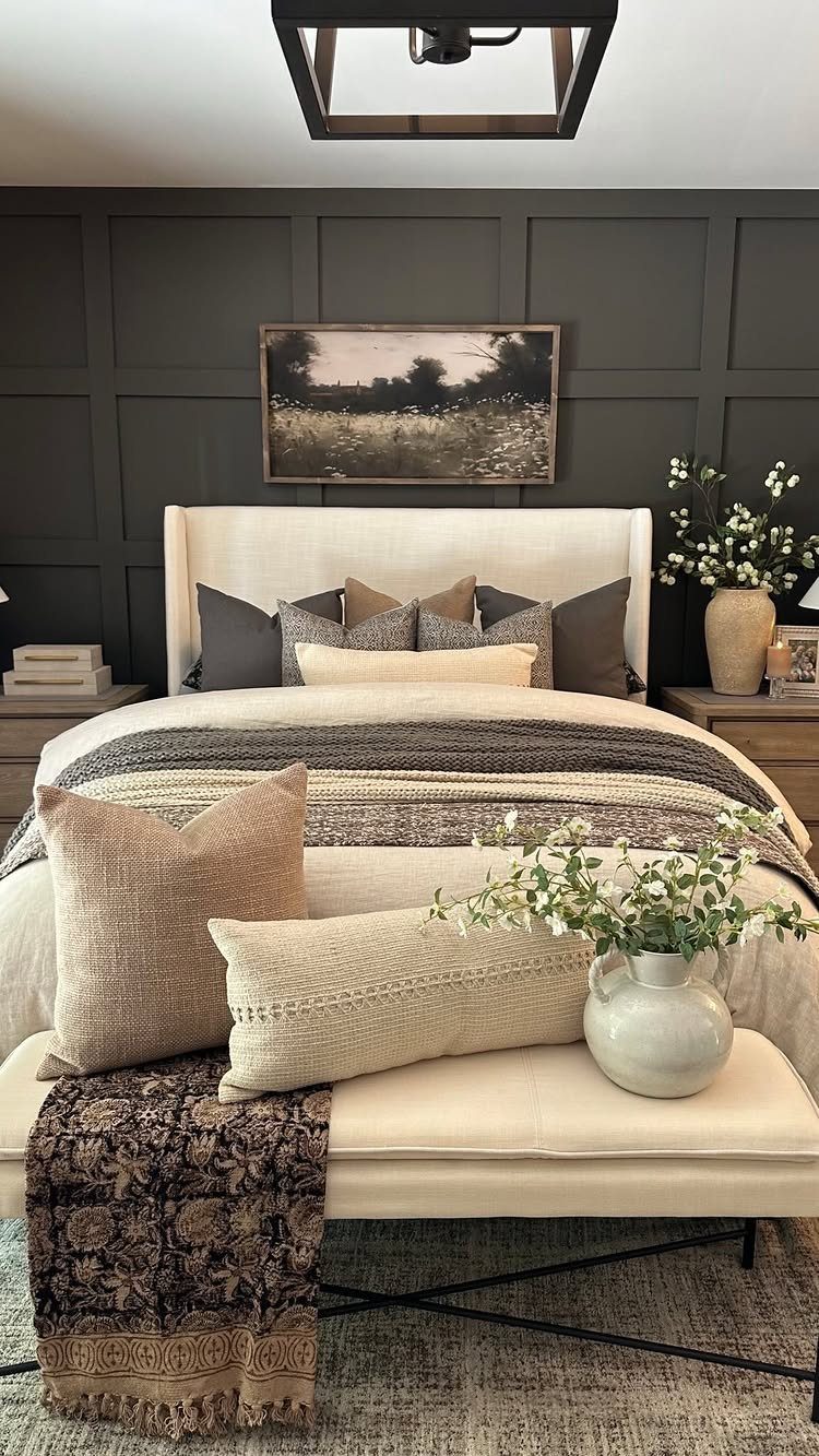Cream bed with layered textured pillows and throws against dark grey paneled walls.