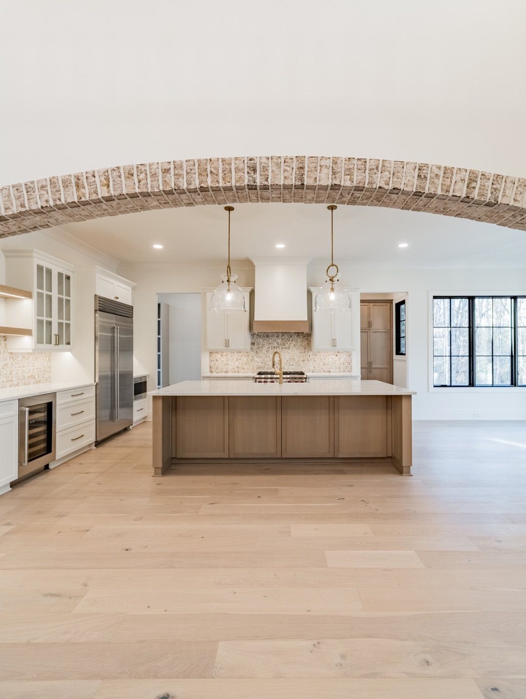 Modern kitchen with light wood floors, white cabinets, and a rustic brick archway.