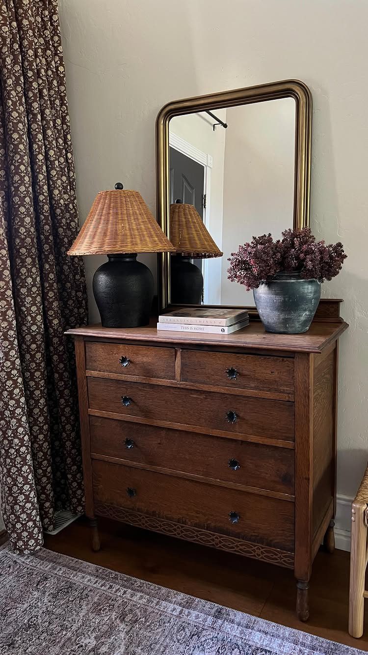 Wooden vintage dresser with a gold-framed mirror, woven lamp, and purple flowers.