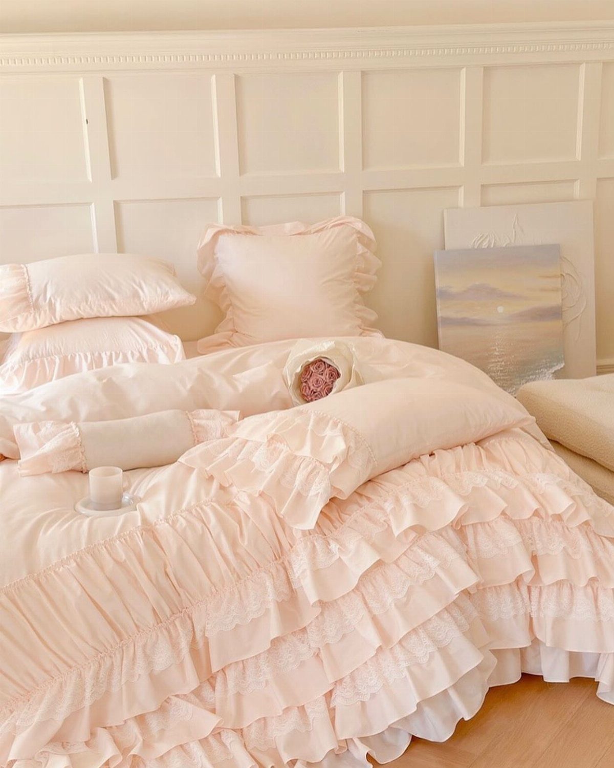 Light pink ruffled bedding and pillows on a bed against white paneled walls.