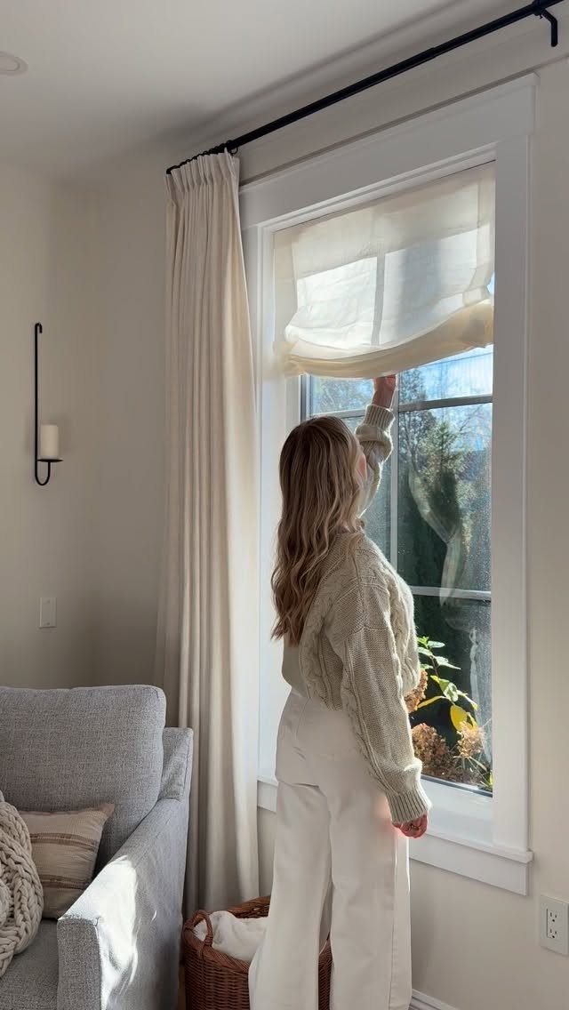 Woman adjusting a white Roman shade and cream curtains in a bright room.