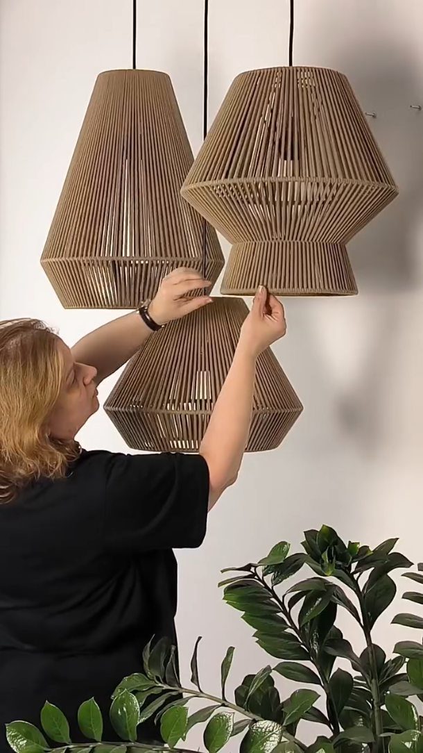 Woman adjusting three large, uniquely shaped wooden slat pendant lampshades against white wall