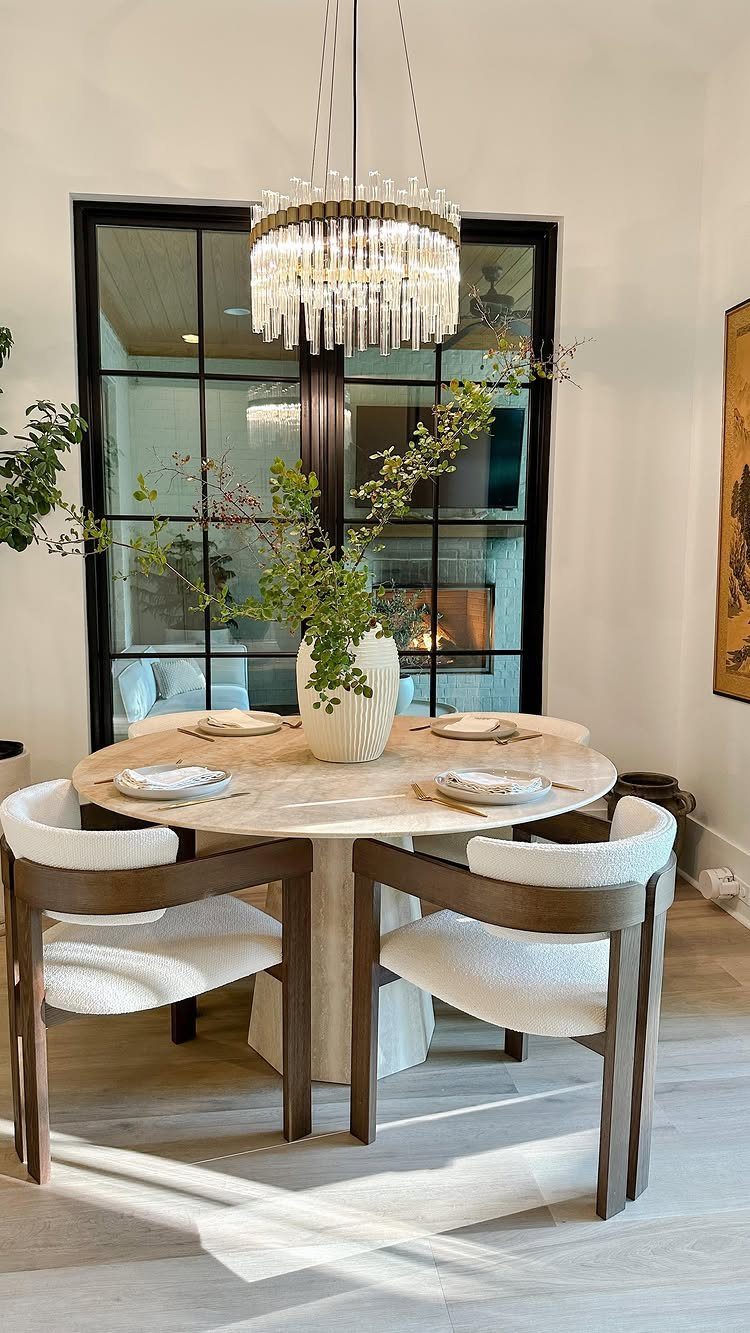 Round marble dining table with white chairs and green branches under a crystal chandelier.