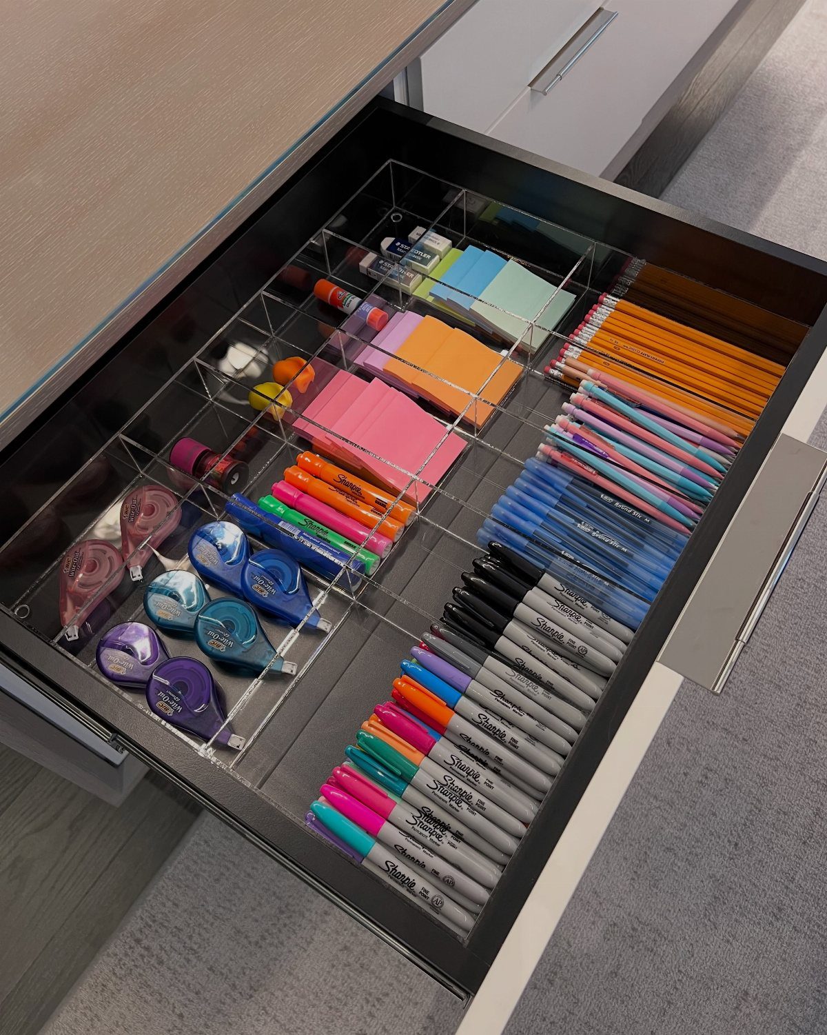 Open desk drawer with clear acrylic organizers holding colorful markers and office supplies.