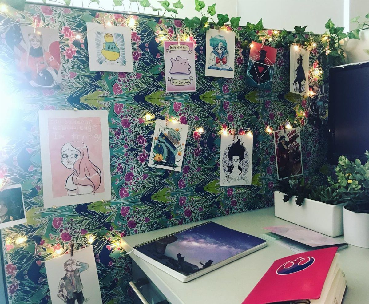 Decorated office cubicle with patterned wallpaper, fairy lights, art prints, and small plants.