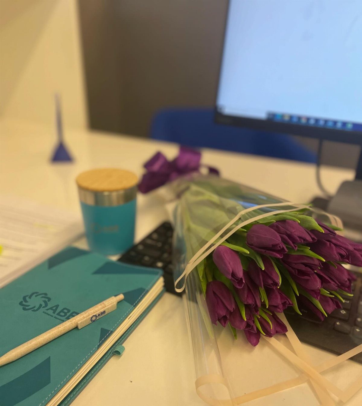 Purple tulips, a teal notebook, and a matching cup on a white office desk.
